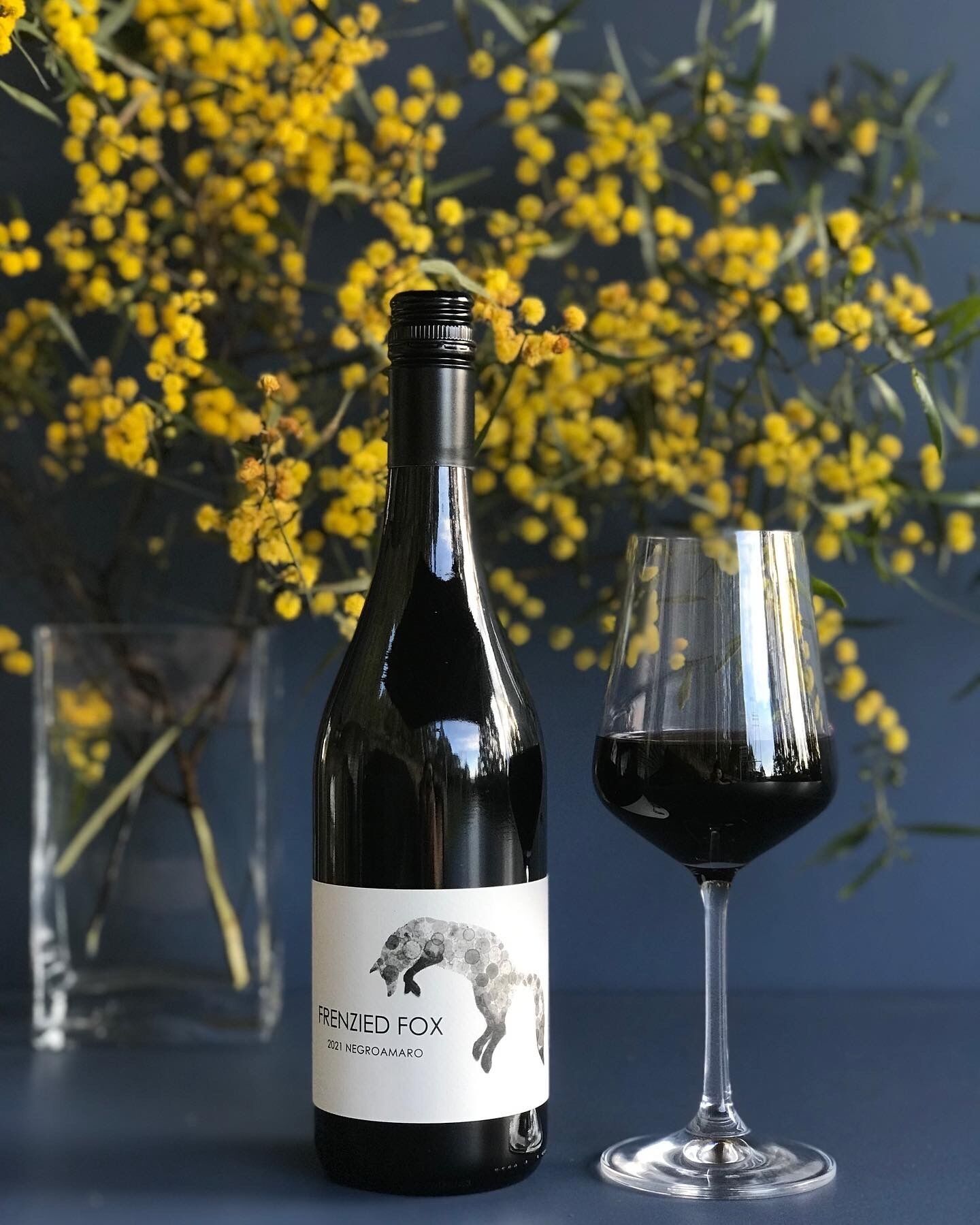 Une bouteille de vin FRENZIED FOX 2021 NEGROAMARO à côté d’un verre à vin, avec un vase de fleurs jaunes en arrière-plan.
