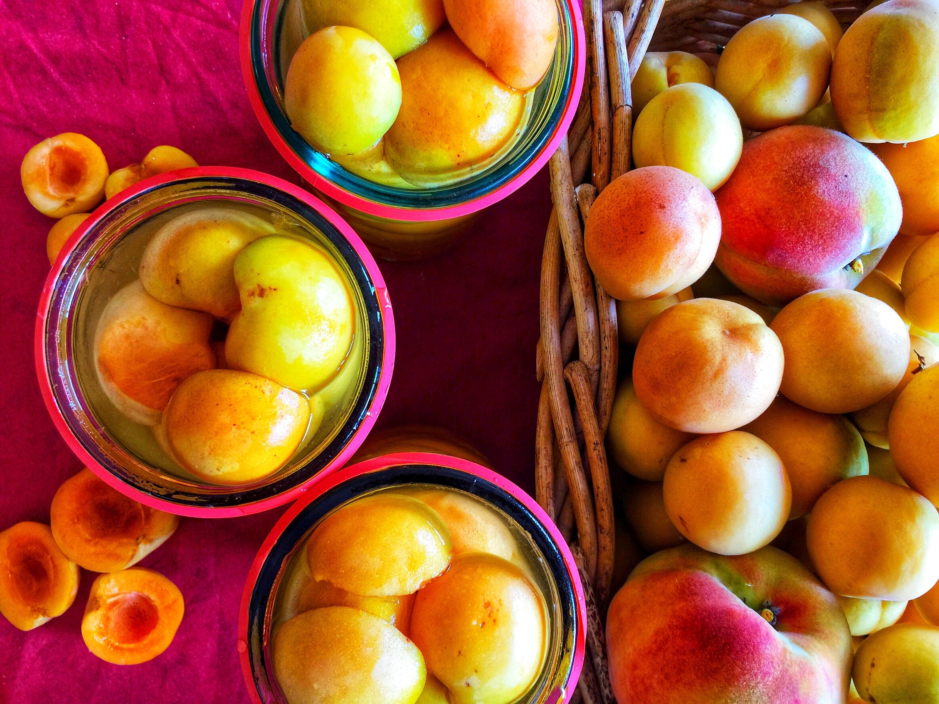 Three glass jars filled with yellow fruits, possibly apricots, on a pink cloth.