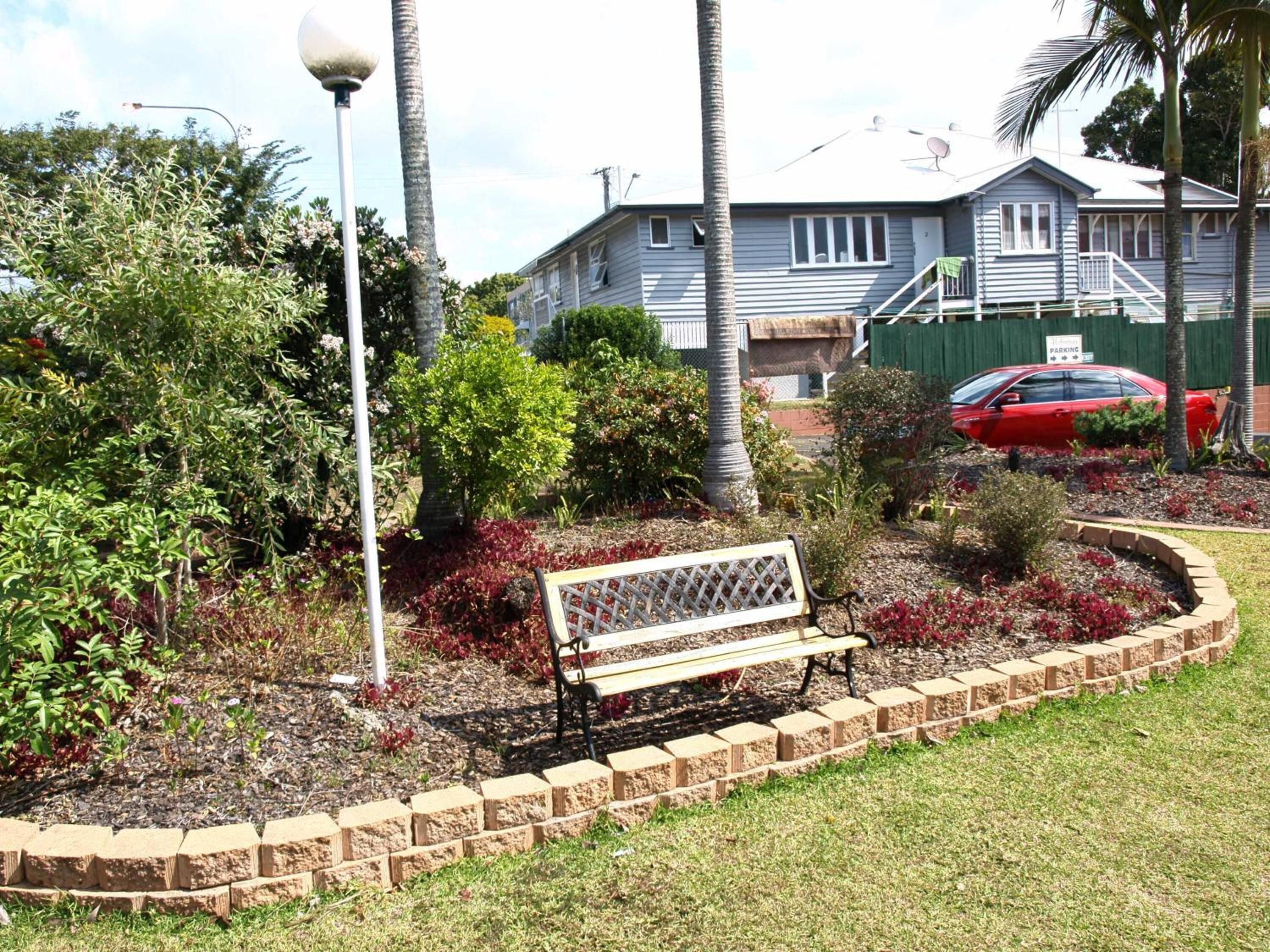 Uma área residencial com um banco em um jardim ajardinado, um carro vermelho estacionado em frente a uma casa e um poste de luz.