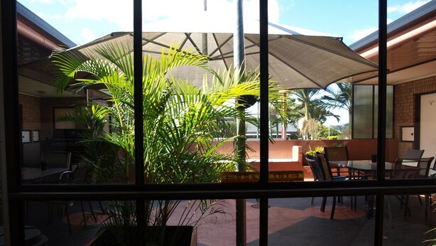 A view through a window of a restaurant with outdoor seating, including a table and chairs, and a large umbrella providing shade. - Lancaster Court Motel (Annerley)