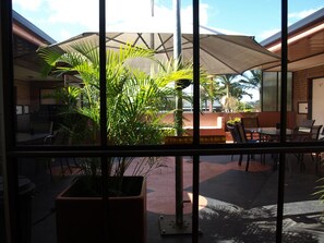 Visão pela janela de um restaurante com mesas ao ar livre, incluindo uma mesa e cadeiras, e um grande guarda-sol para dar sombra.