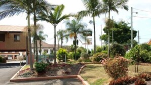 A parking lot with a landscaped area featuring various plants and a building in the background. - Lancaster Court Motel (Annerley)