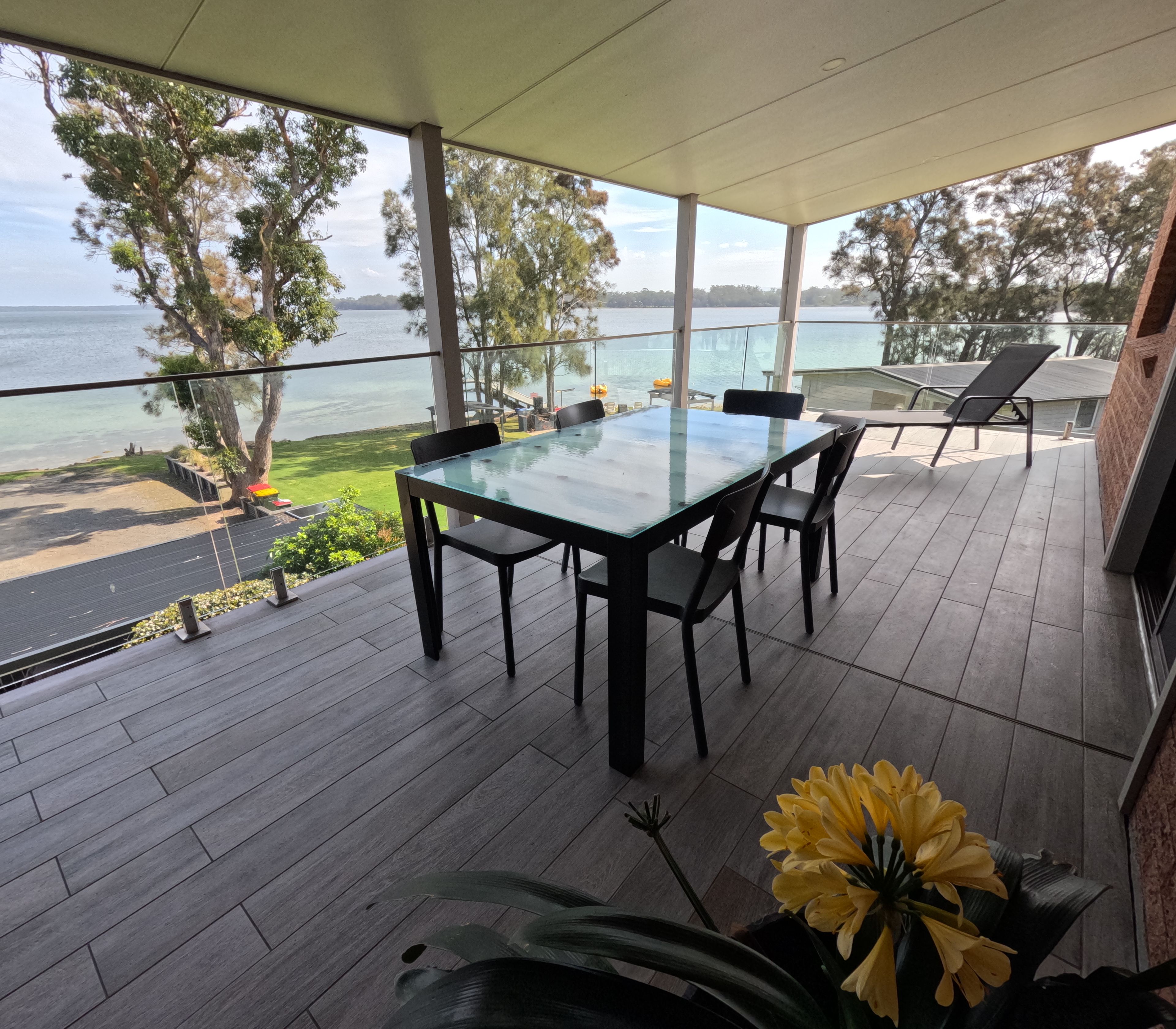 A covered outdoor area with a glass table and chairs, overlooking a body of water and greenery.