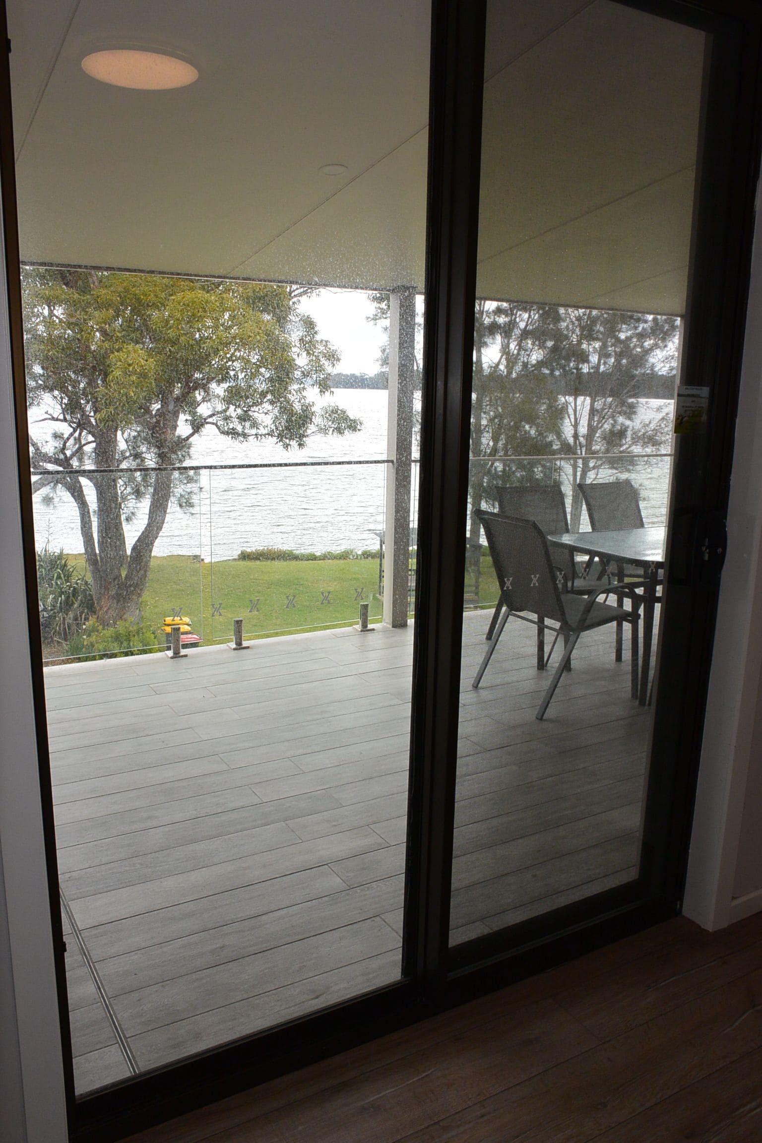 A view through a glass door of a deck with a table and chairs, overlooking a body of water and a grassy area with trees.