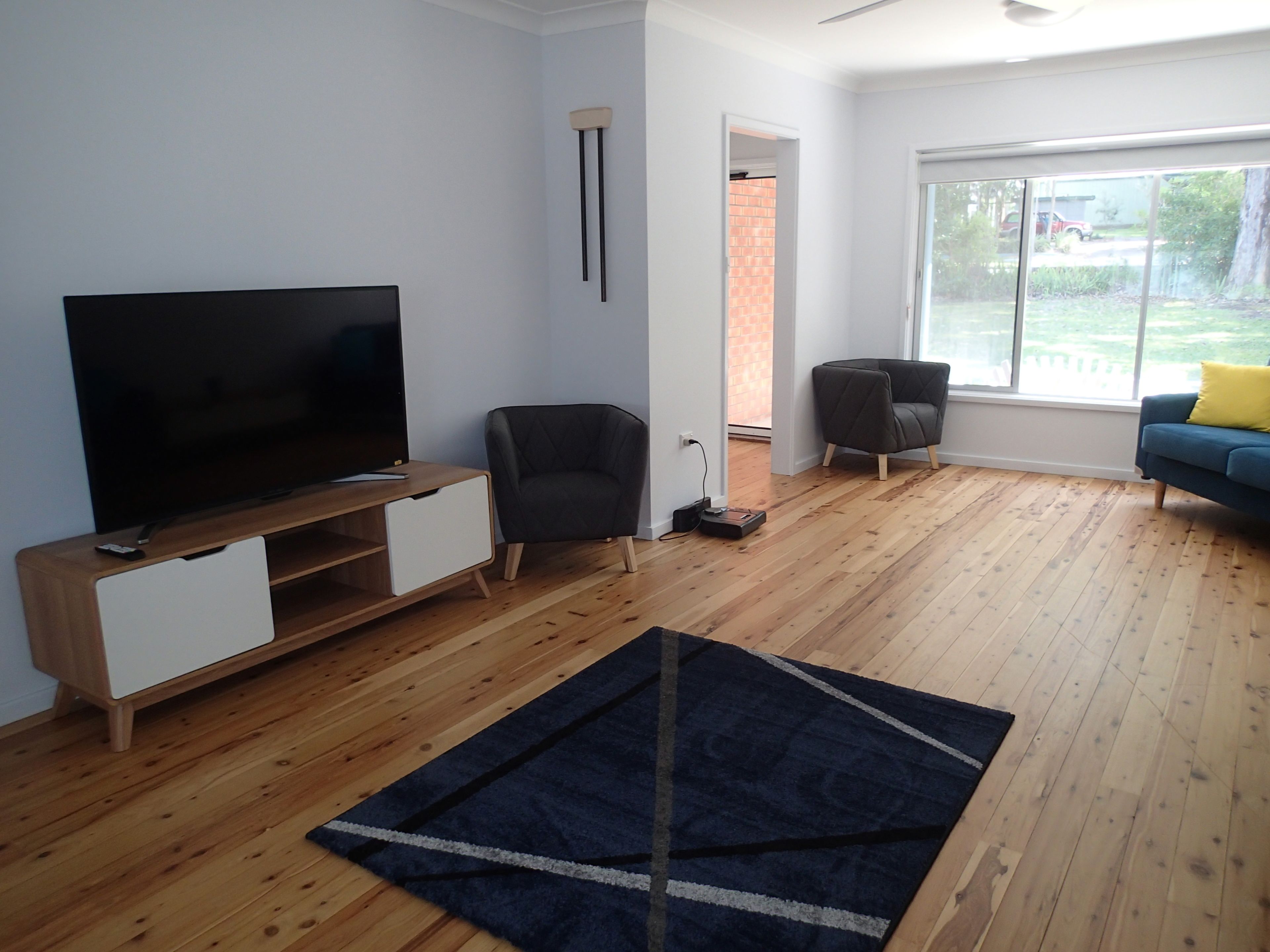 A modern living room with a flat-screen TV, a dark armchair, a blue sofa with a yellow pillow, and a large window offering a view of the outdoors.