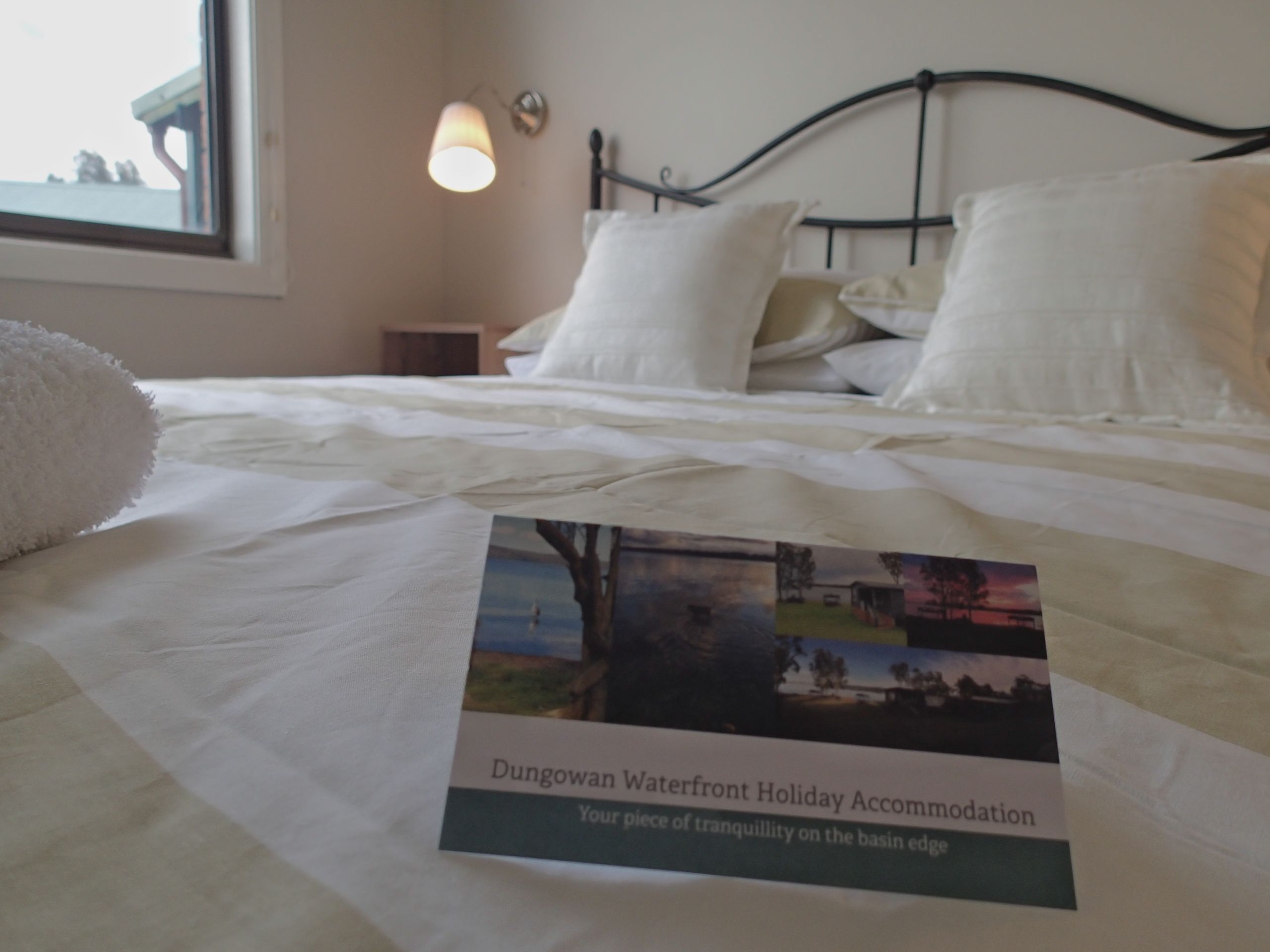 A neatly made bed with white linens and pillows, a framed picture of a scenic landscape, and a small bedside table with a lamp.