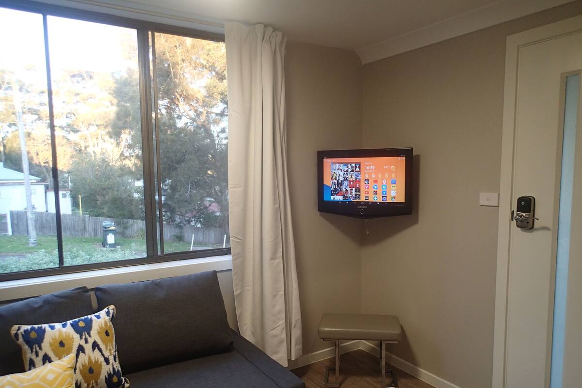 Ein Wohnzimmer mit grauem Sofa, einer kleinen Bank, einem an der Wand montierten Fernseher und einem Fenster mit Vorhängen.