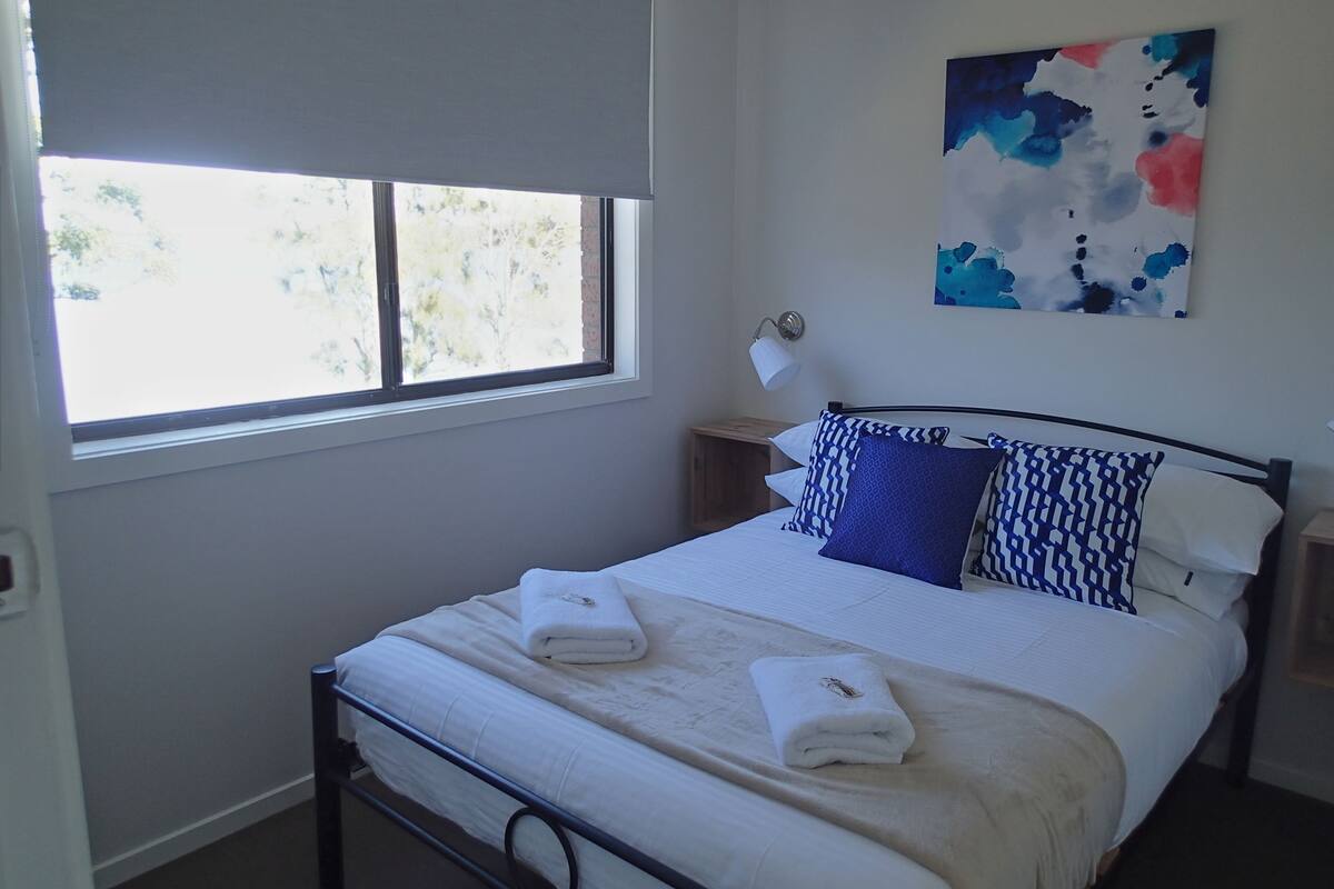 Ein ordentlich bezogenes Bett mit weißen Leinentüchern und zwei Handtüchern, ein Fenster mit Blick auf Bäume, ein an der Wand hängendes abstraktes Gemälde und eine Nachttischlampe.
