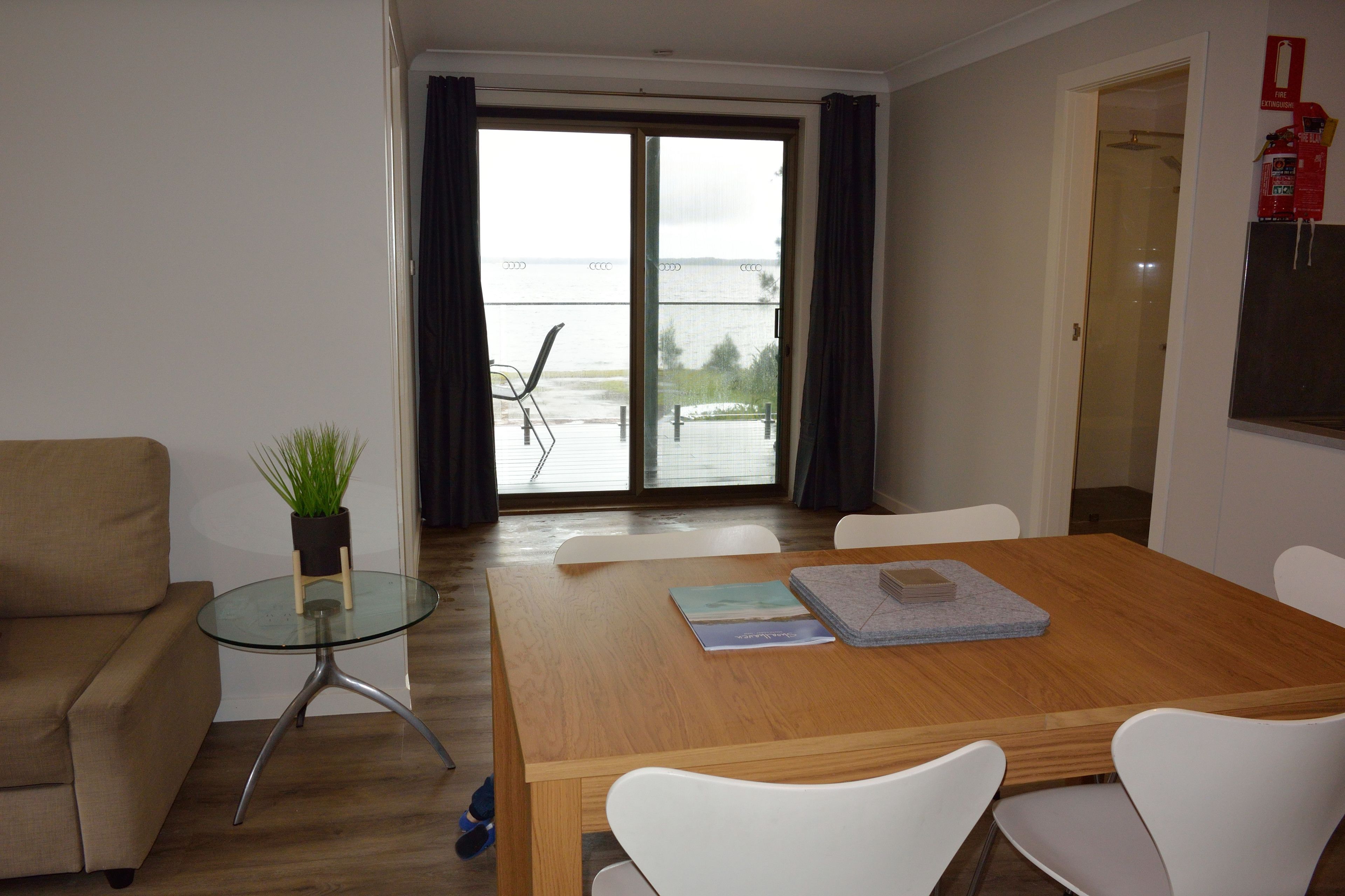 A modern living room with a wooden dining table, white chairs, a glass coffee table, and a beige sofa. There is a view of a body of water through a glass door.