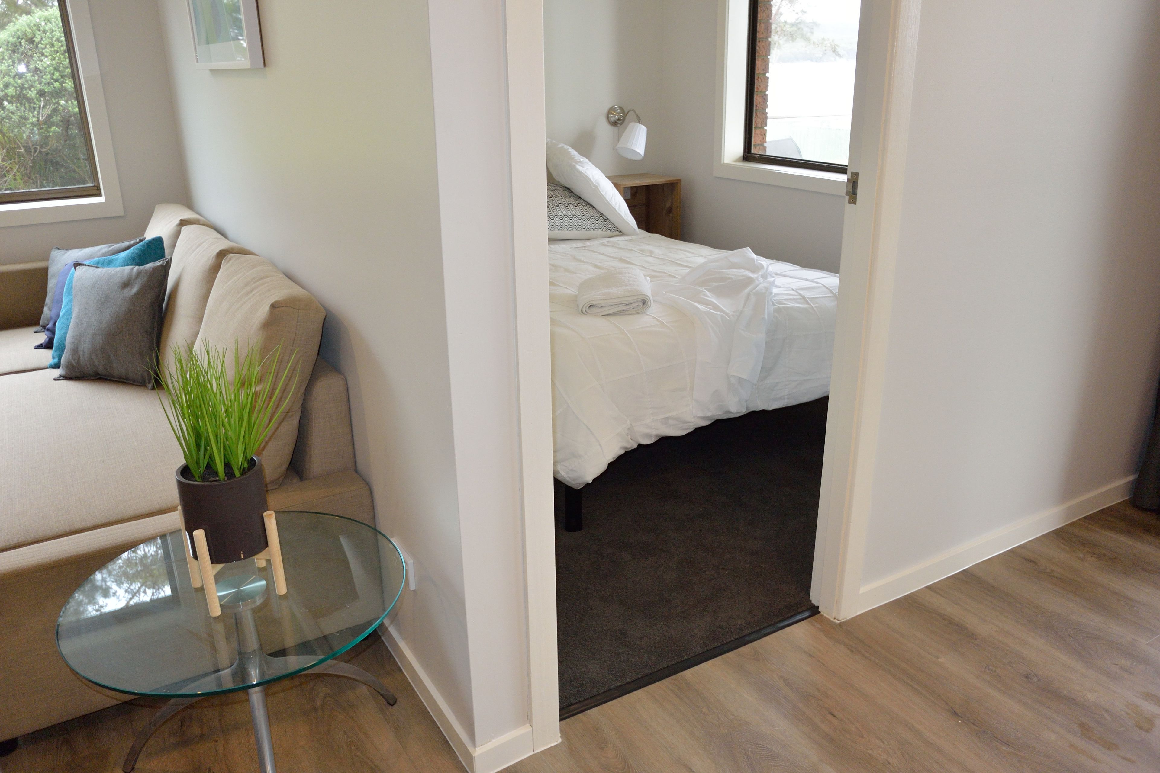 A bedroom with a bed, a glass table with a plant, a sofa with cushions, and a window.