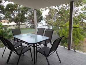 A covered outdoor area with a glass table and chairs, surrounded by a railing with a view of trees and a distant building. - Dungowan Waterfront Apartments (Erowal Bay)