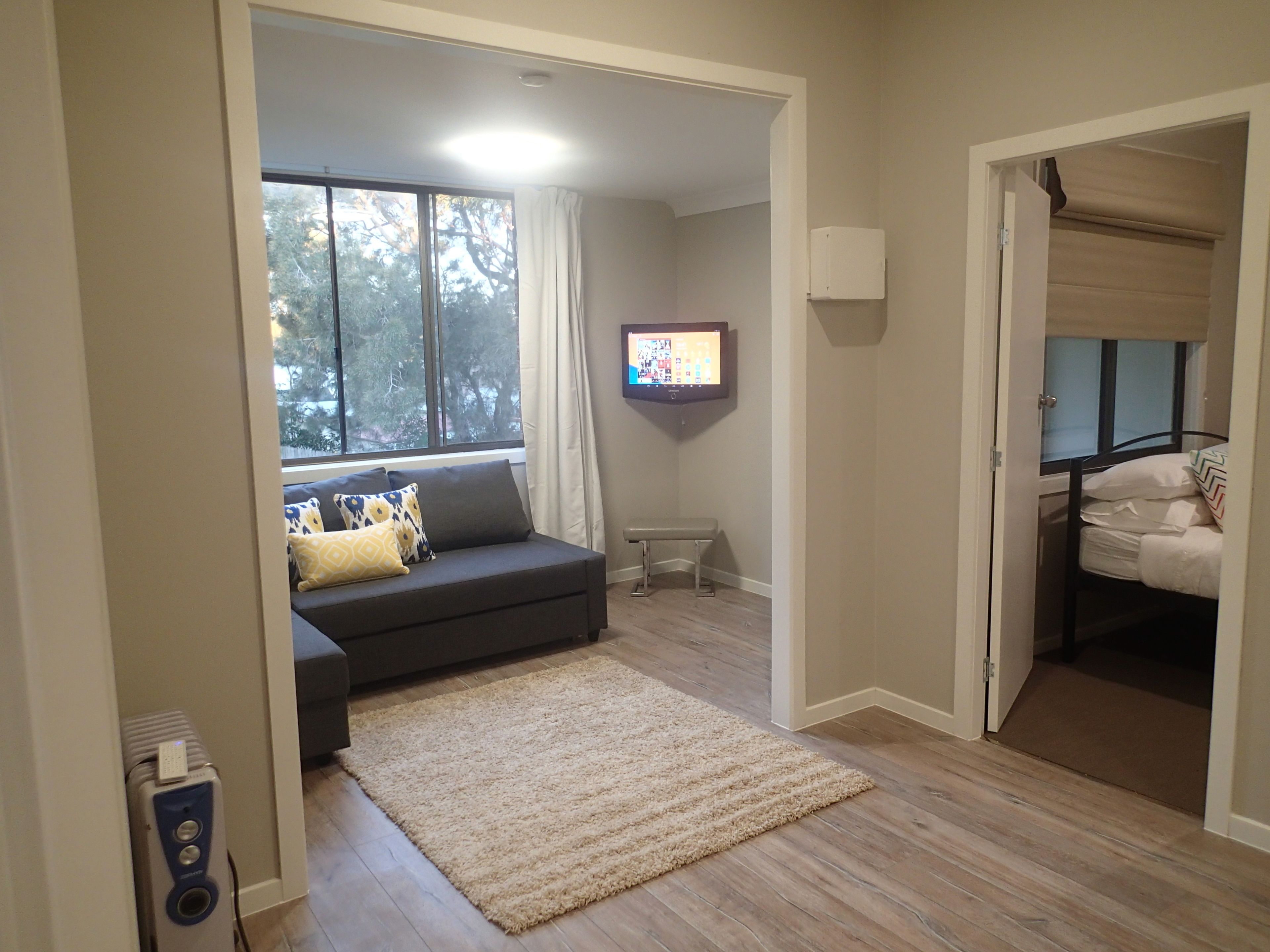 A living room with a grey sofa, a beige rug, a TV on the wall, and a window with curtains.
