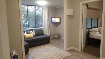 A living room with a grey sofa, a beige rug, a TV on the wall, and a window with curtains.
