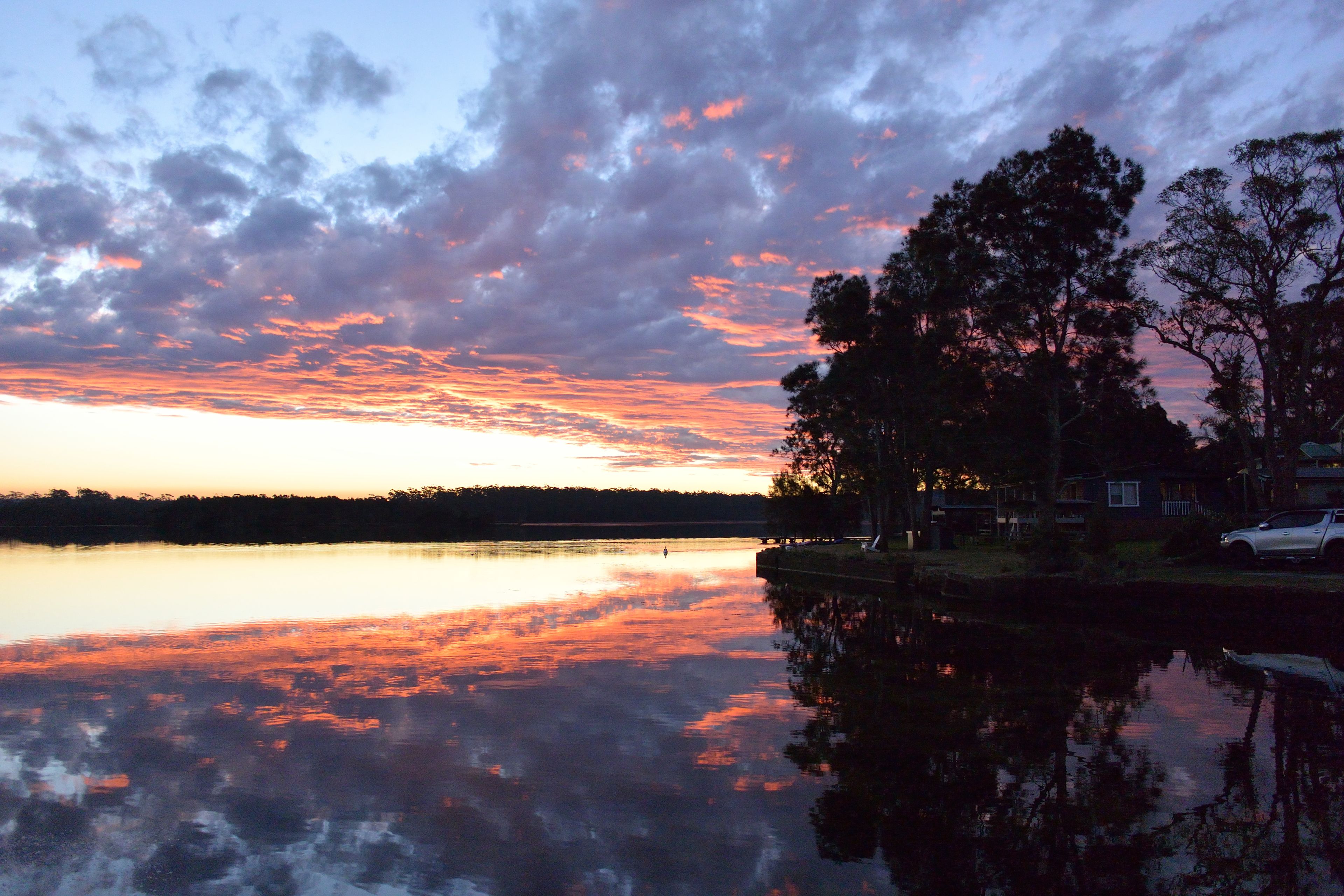 A calm lake at sunset with a reflection of the sky, surrounded by trees and a residential area.