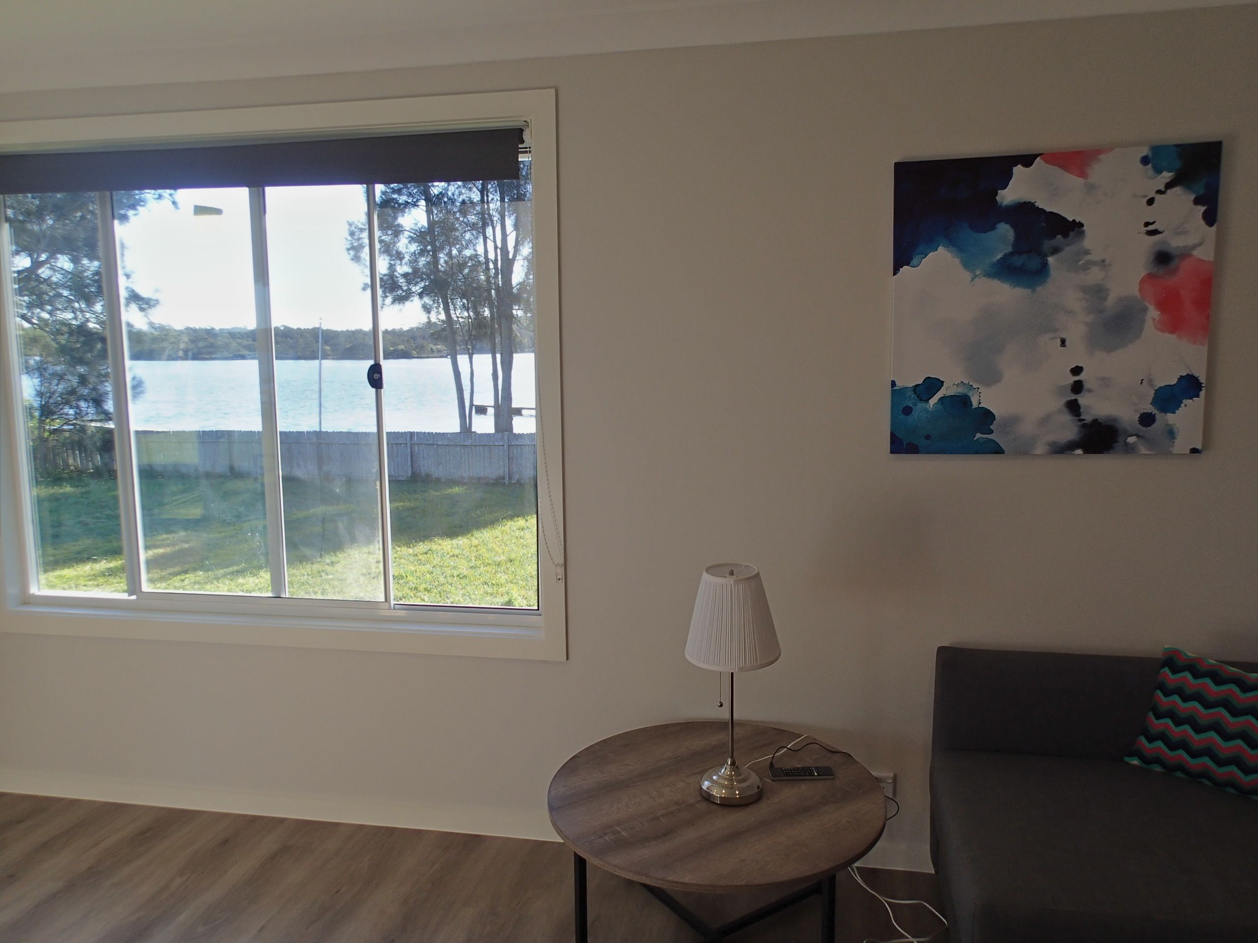 Una habitación con un ventanal grande que ofrece vistas a un lago y árboles, una mesa redonda de madera con una lámpara, un sofá gris y un cuadro abstracto colorido en la pared.