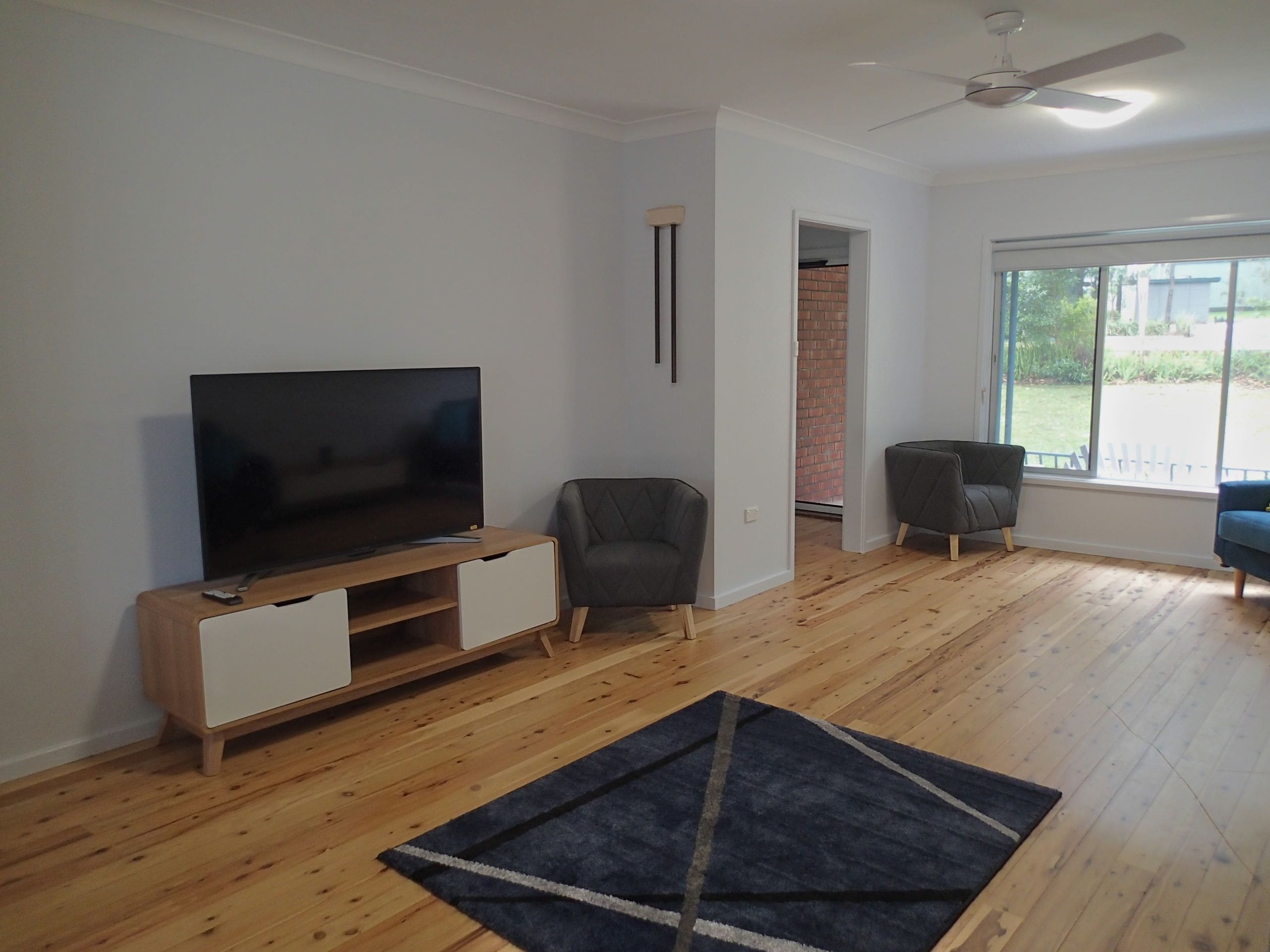 A living room with a flat-screen TV on a wooden TV stand, a grey armchair, a dark rug, and a large window with a view of greenery.