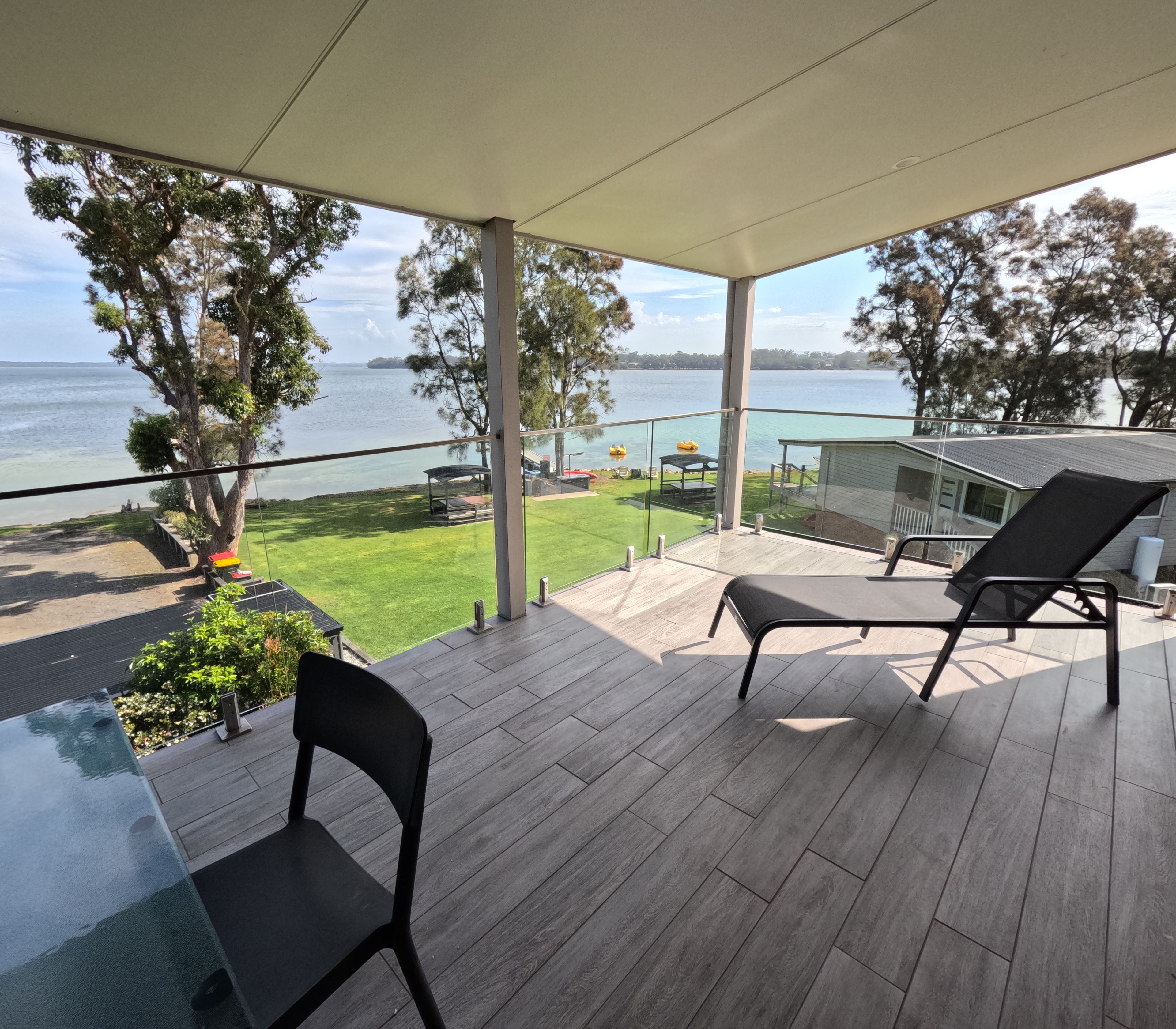 A covered outdoor area with a view of the water, a table with chairs, and a grassy lawn.