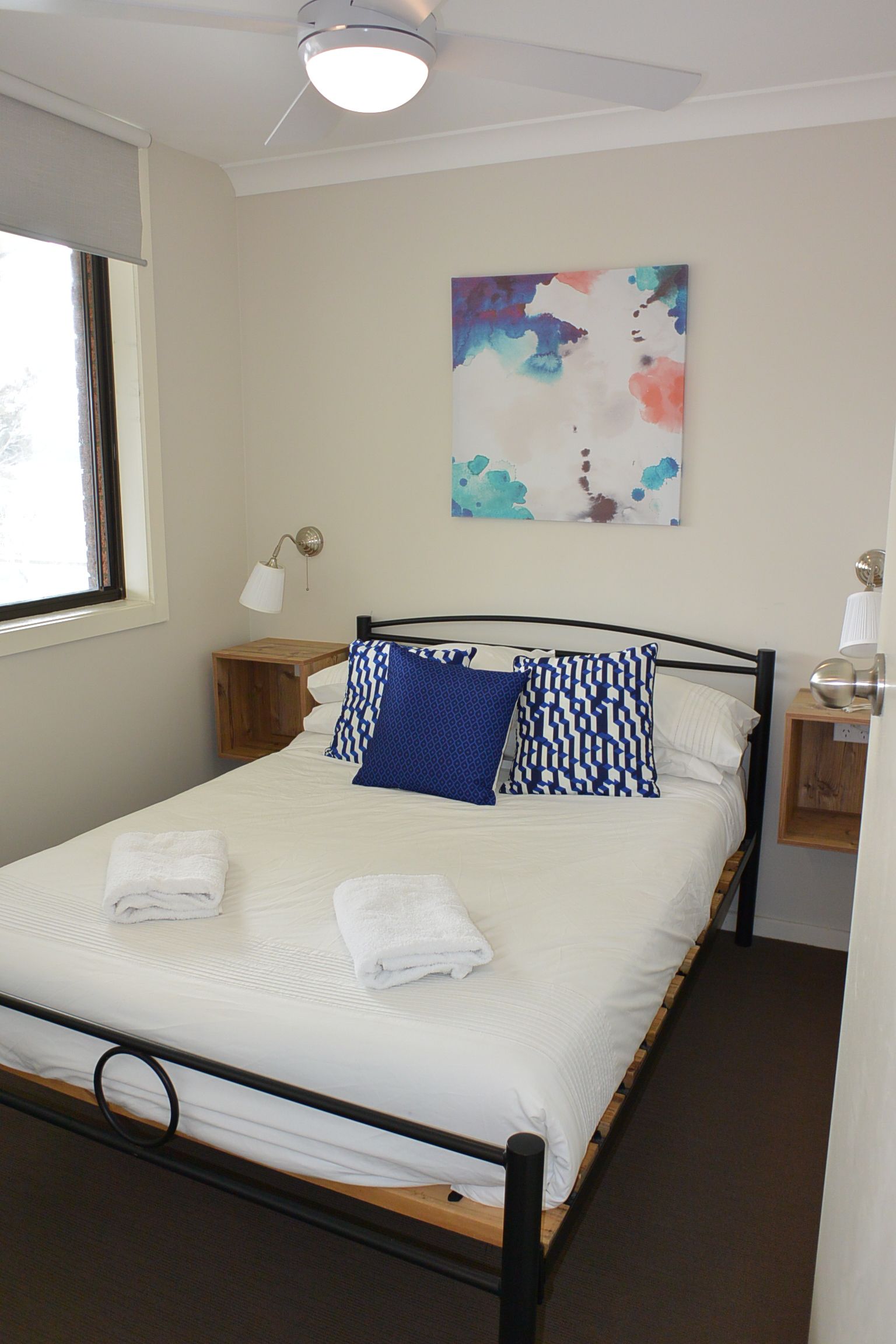 A neatly made bed with white linens and blue patterned pillows, a ceiling fan, and a wall-mounted abstract painting.
