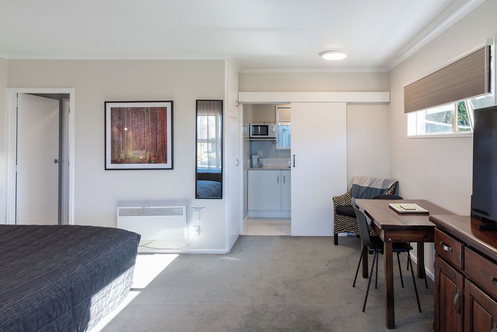 Een slaapkamer met een bed, een bureau en een stoel, een kitchenette op de achtergrond en een raam met jaloezieën.