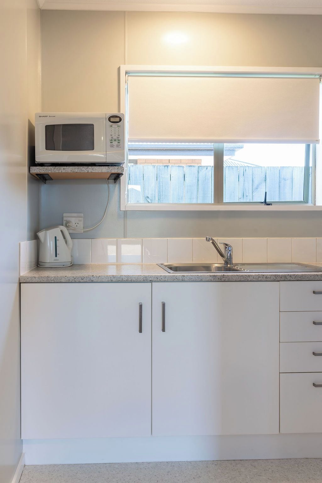 Una cocina con armarios blancos, microondas, hervidor y una ventana sobre el fregadero.