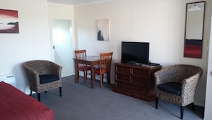 A room with a bed, two wicker chairs, a wooden table with chairs, a TV on a wooden cabinet, and artwork on the walls. - Mount Hutt Motels (Methven)