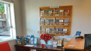 A reception area with a wooden bookshelf displaying DVDs, a red chair, a desk with a computer, and a small table with a vase of red flowers. - Mount Hutt Motels (Methven)