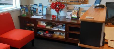 Zona de recepción con una estantería de madera que exhibe DVDs, una silla roja, un escritorio con una computadora y una pequeña mesa con un jarrón de flores rojas.