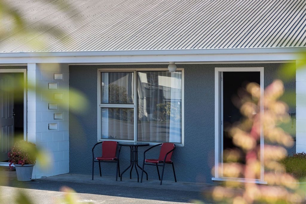 Un edificio con fachada gris, una ventana con cortina y dos sillas rojas afuera.