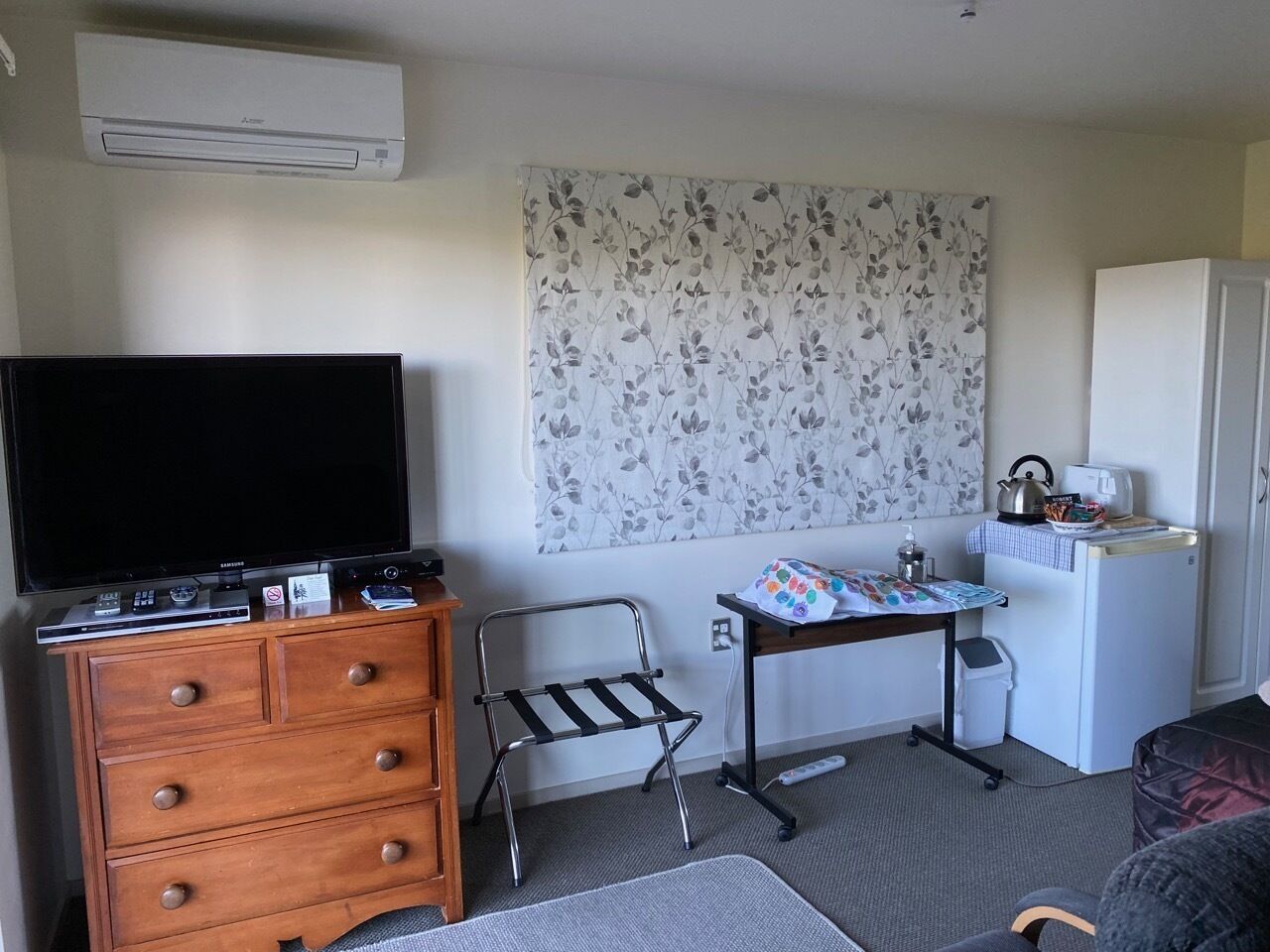 Habitación de hotel compacta con televisor de pantalla plana, cómoda de madera, pequeña cocina y pared con estampado floral.