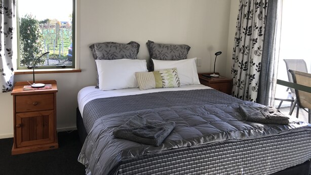A neatly made bed with a grey quilt, two white pillows, and a grey throw blanket. There is a wooden nightstand with a lamp and a small table beside the bed. A window with curtains is visible, and a chair is placed near the window. - Bluebell Lodge And Cottage (Hastings)