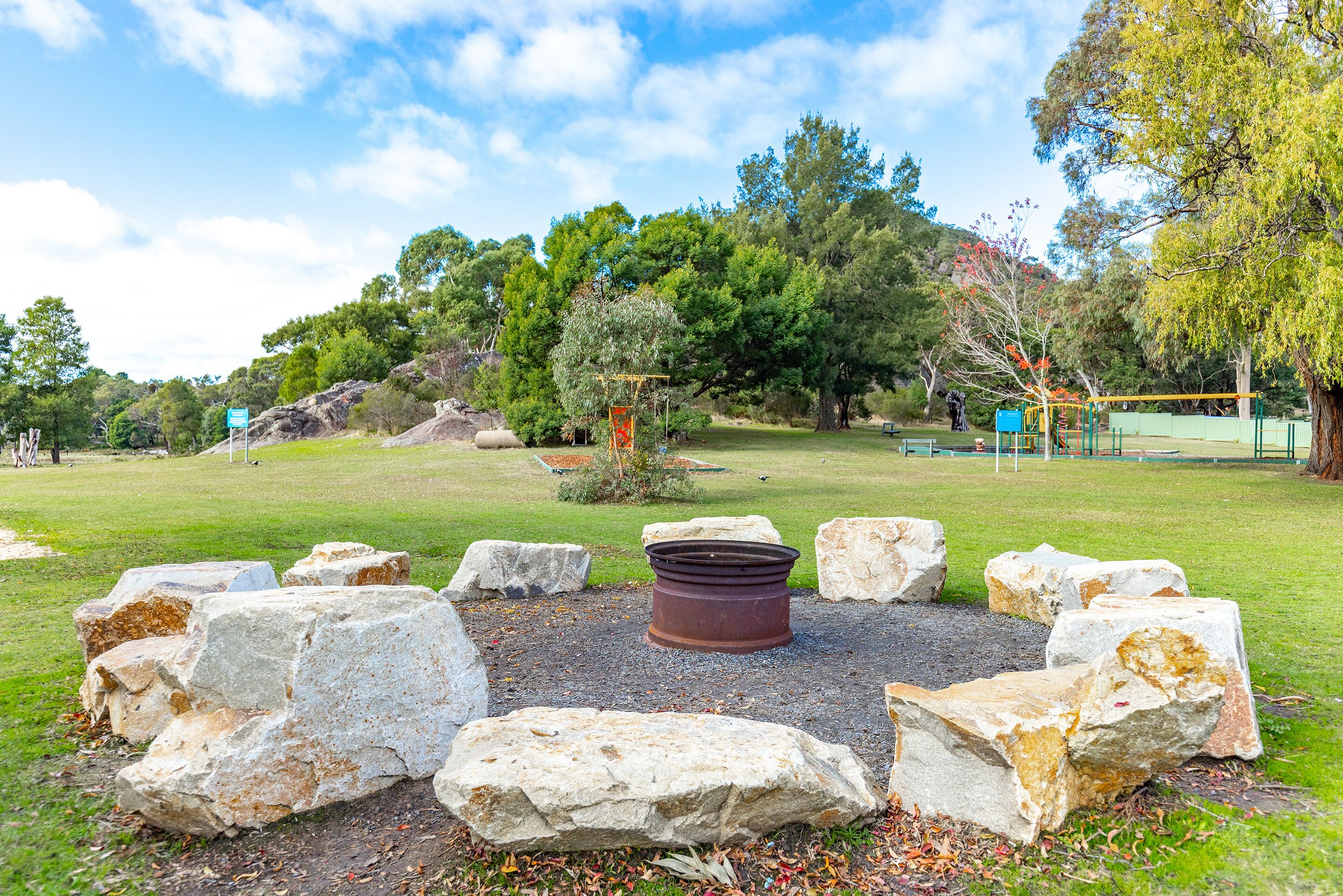 A park with a central fire pit surrounded by large rocks, trees, and a playground in the background.