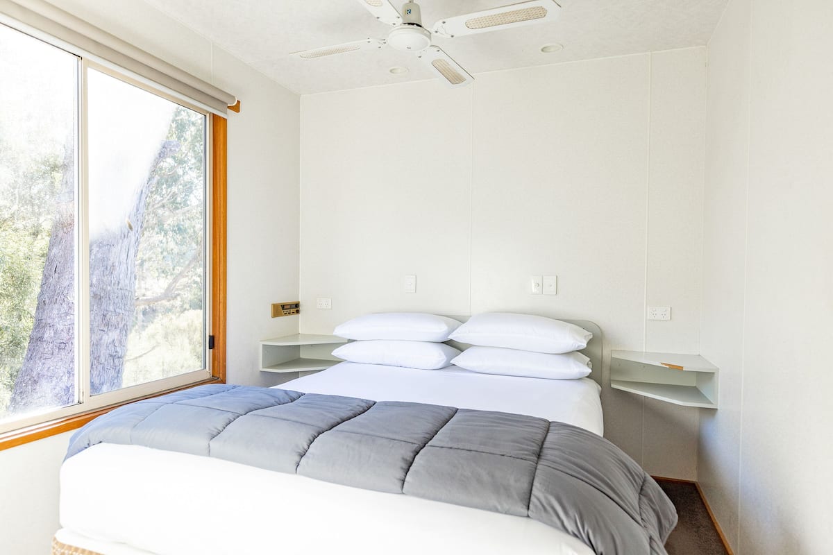 A bedroom with a bed, white walls, a ceiling fan, and a window with a view of trees.