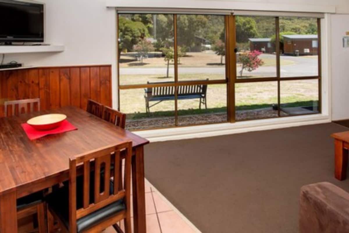 A room with a wooden dining table, a bench outside the window, and a television mounted on the wall.