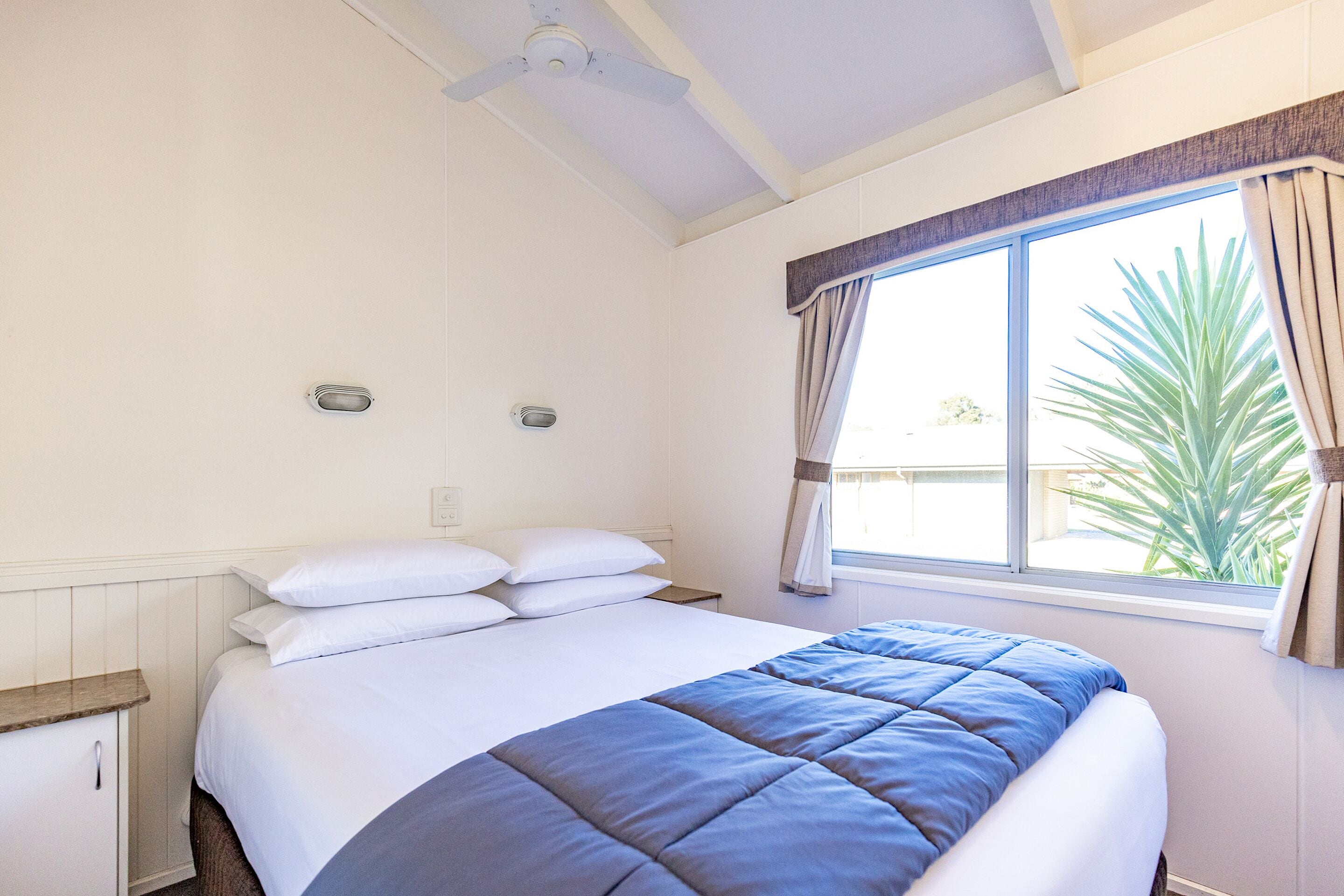 A bedroom with a bed, a window with curtains, a ceiling fan, and a small bedside table.