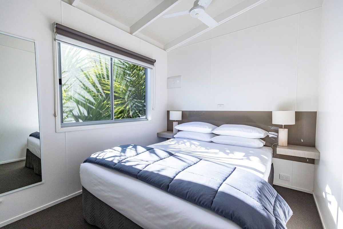 A bedroom with a bed, a nightstand, a lamp, a window with blinds, and a ceiling fan.