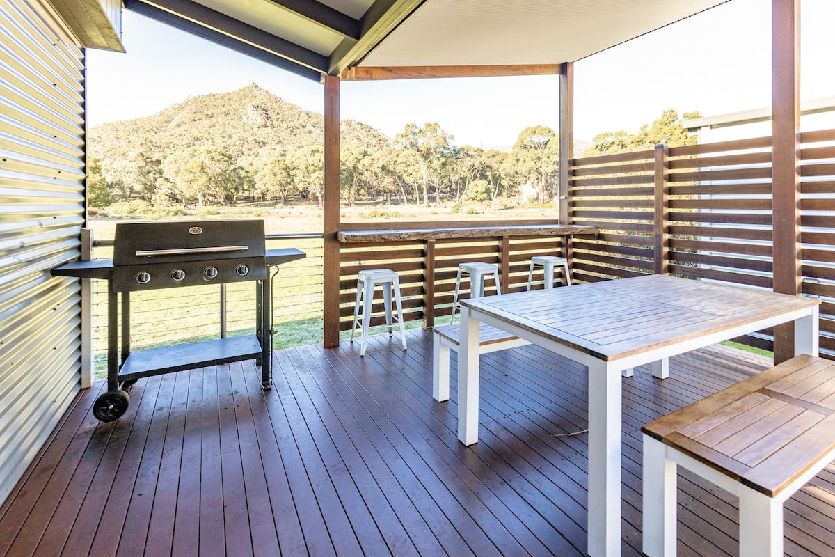 A covered outdoor area with a wooden deck, a barbecue grill, white tables and benches, and a view of a hilly landscape.