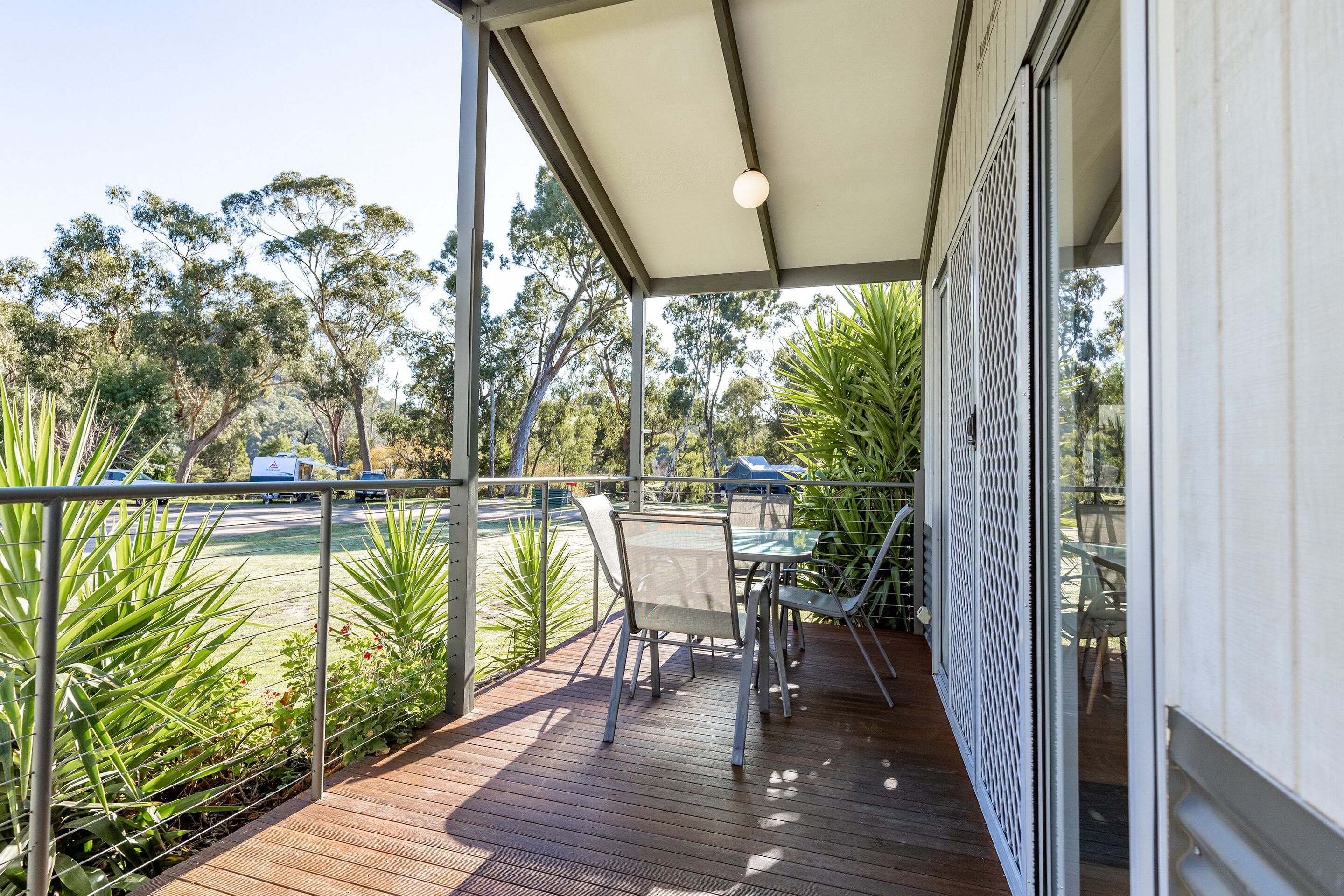 A covered outdoor area with a table and chairs, surrounded by greenery and a view of a road.