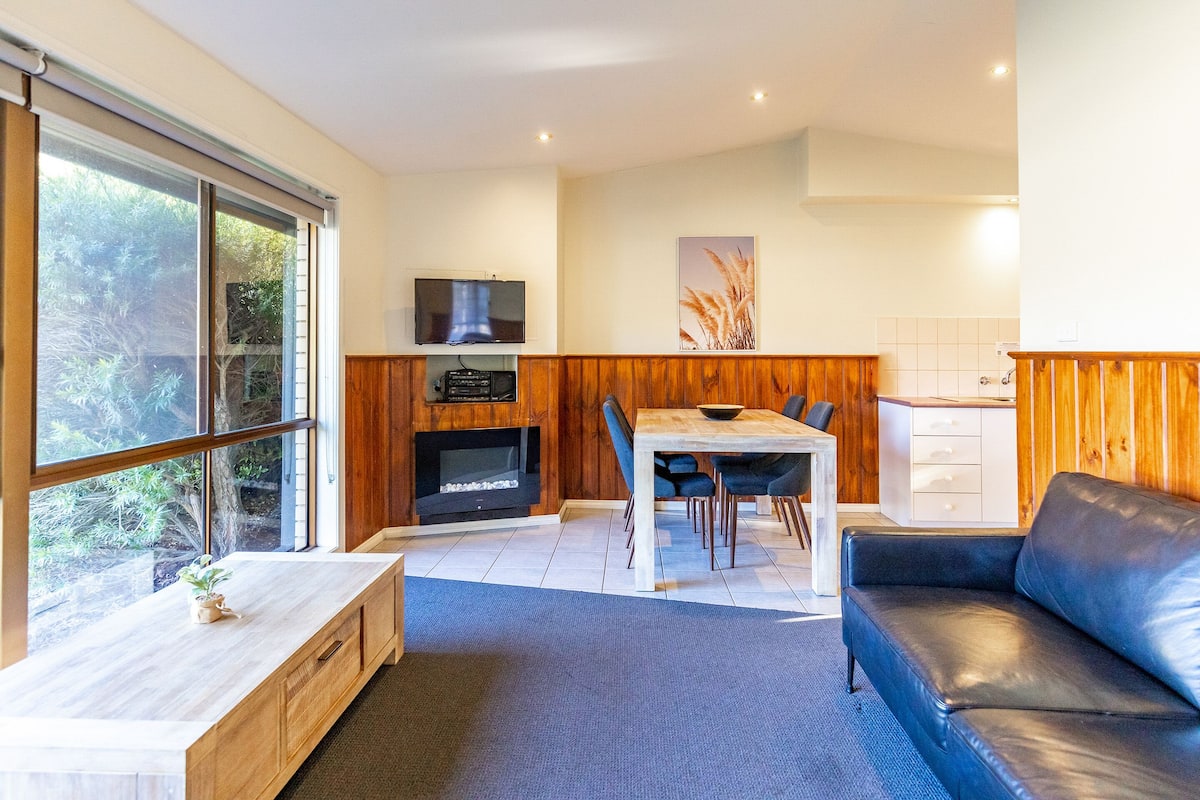 A modern living room with a fireplace, wooden paneling, a dining area, and a black leather sofa.