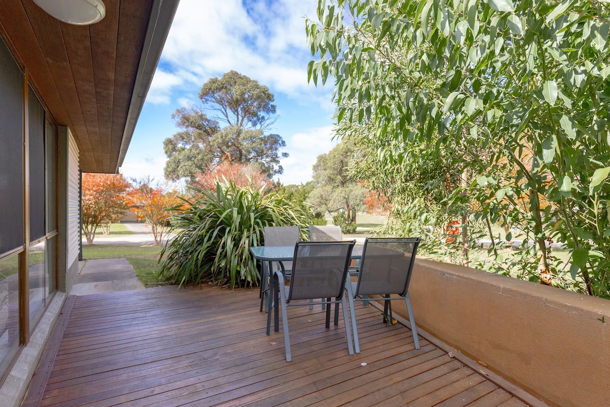 A wooden deck with a table and chairs, surrounded by greenery and trees.