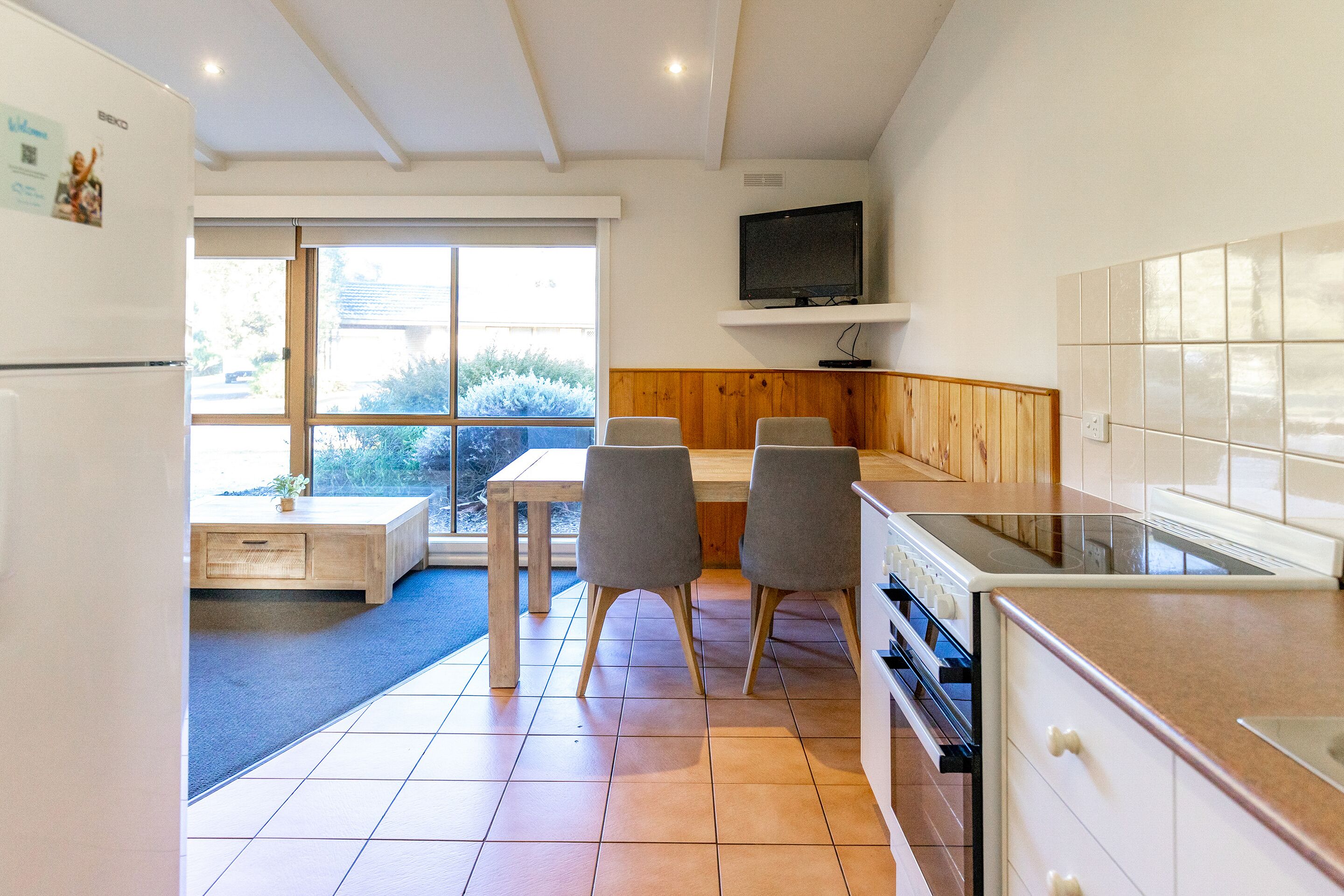 A modern kitchen with a dining area, wooden paneling, a TV mounted above the counter, and a large window providing a view of the outdoors.