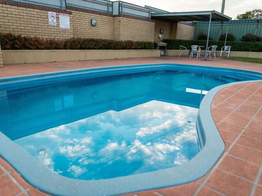 Piscina com reflexo do céu límpido na água, rodeada por um pátio de tijolos e um muro de tijolos com uma placa.