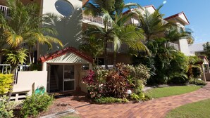 A modern multi-story building with a central entrance, surrounded by lush greenery and palm trees.