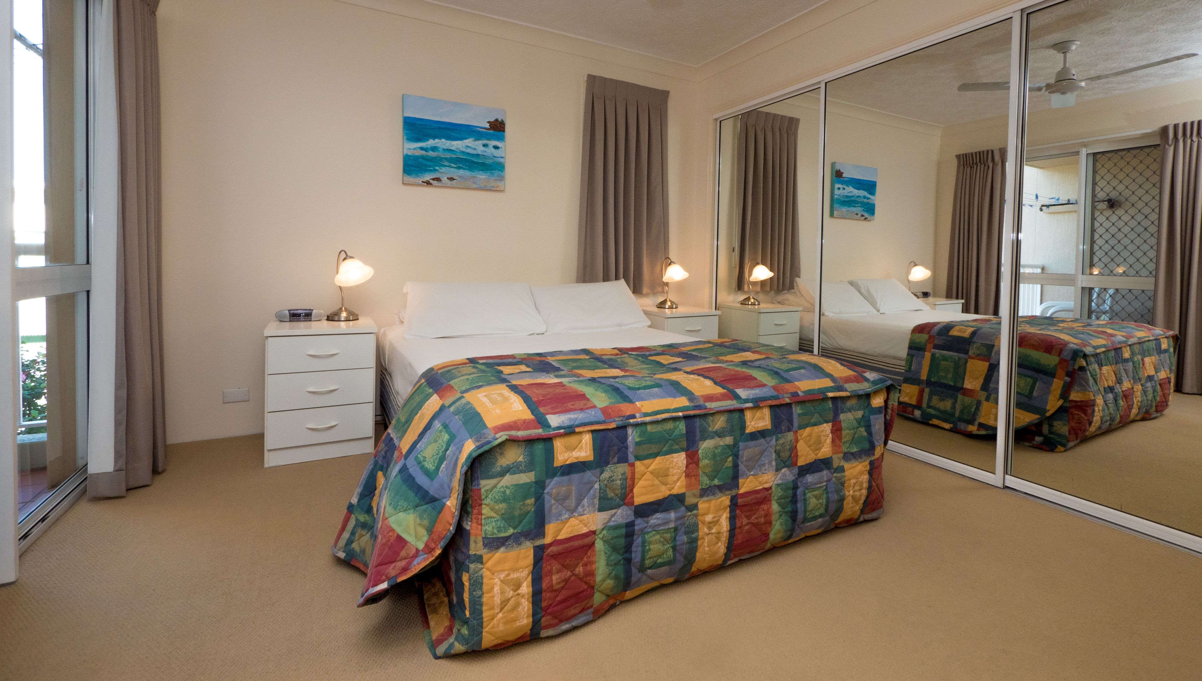 A bedroom with a large mirror, a bed with a colorful quilt, two bedside tables with lamps, and a painting on the wall.