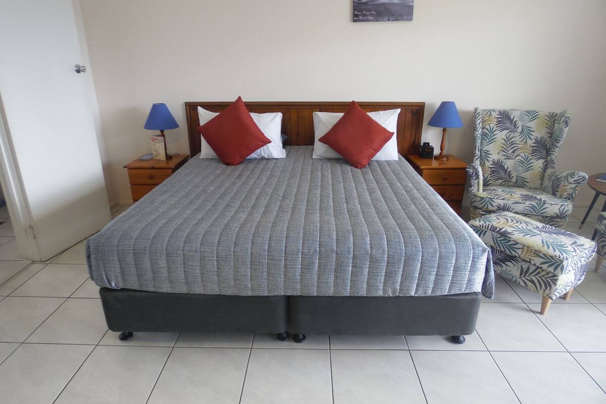 Una cama bien hecha con colcha gris, dos almohadas rojas y un cabecero de madera. Hay dos mesitas de noche con lámparas, una silla con estampado de hojas y un cuadro en la pared.