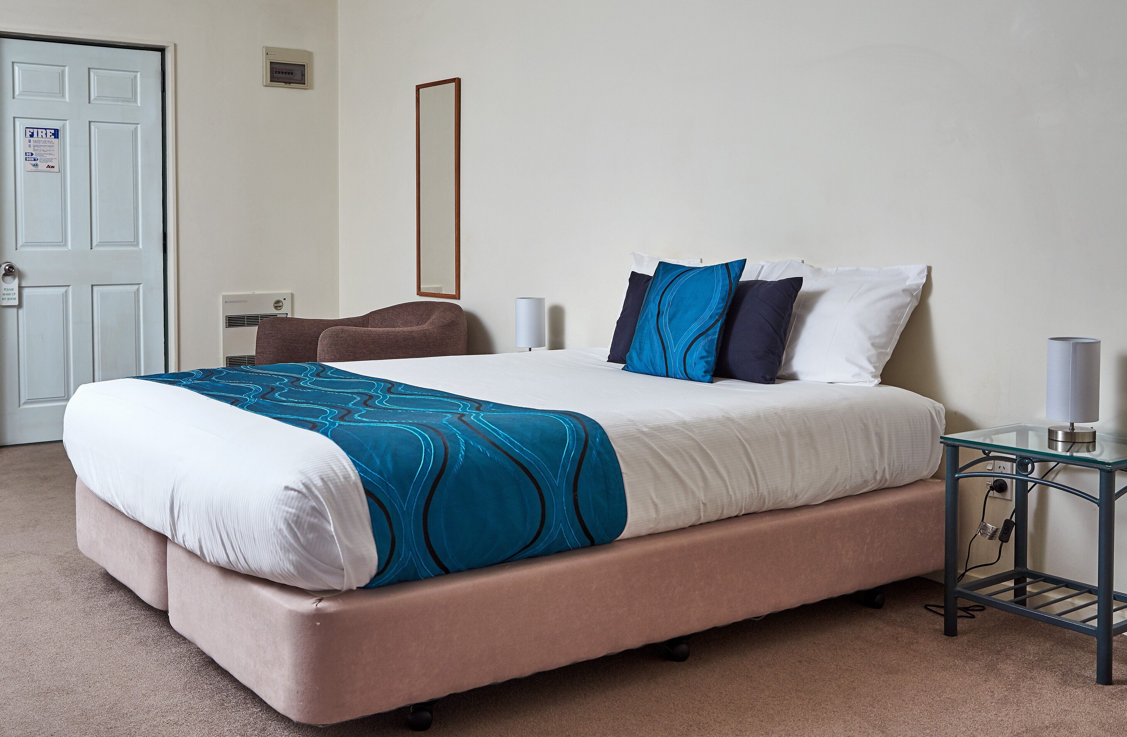 A neatly made bed with a blue and white bedspread, a brown chair, a bedside table with a lamp, and a door with a fire safety sign.