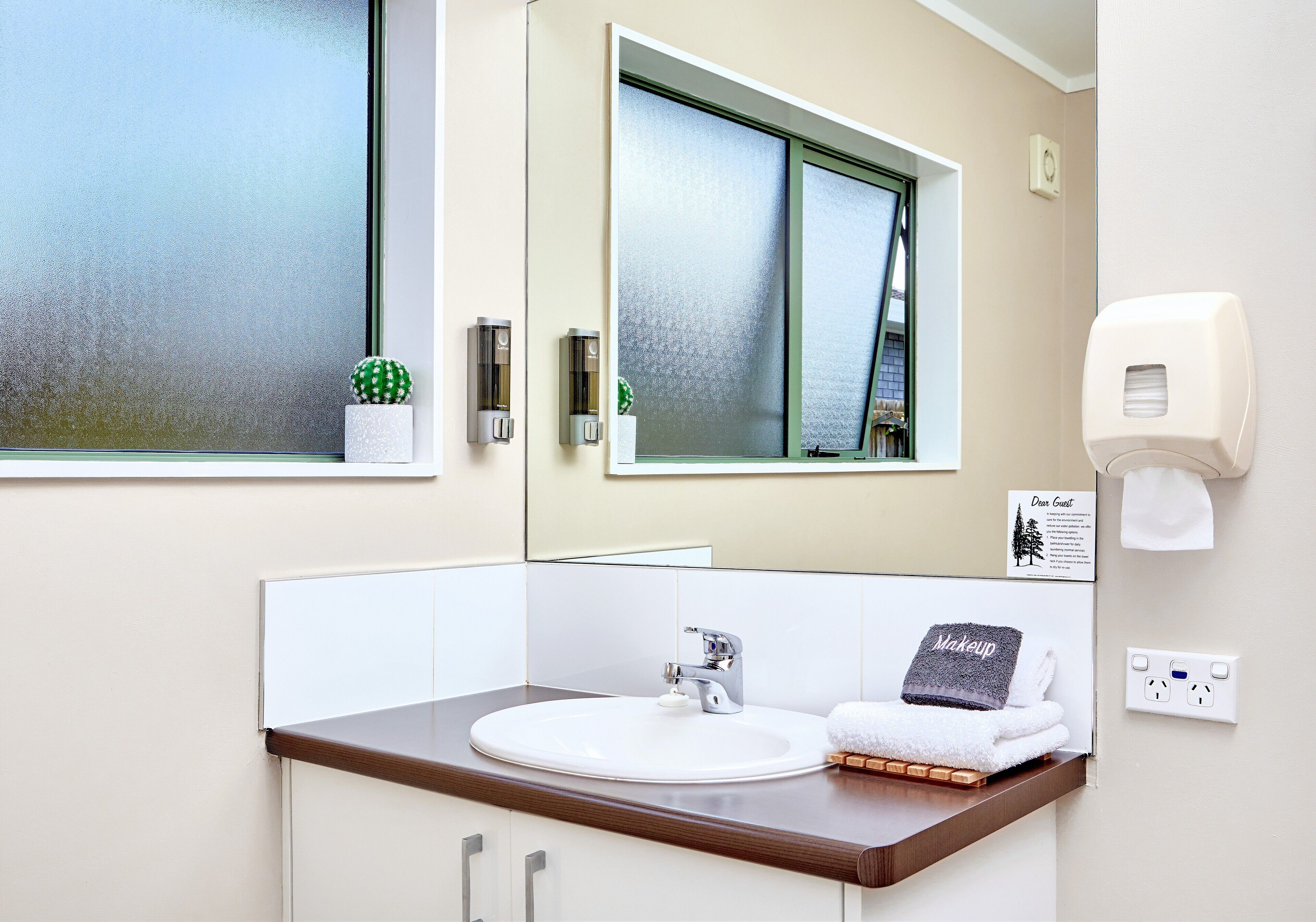 A bathroom with a white sink, a mirror, a window, a towel rack, and a soap dispenser.