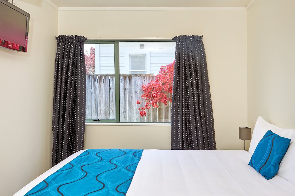 Un dormitorio con cama, ventana con cortinas, televisor y vista a una cerca de madera y flores rojas afuera.
