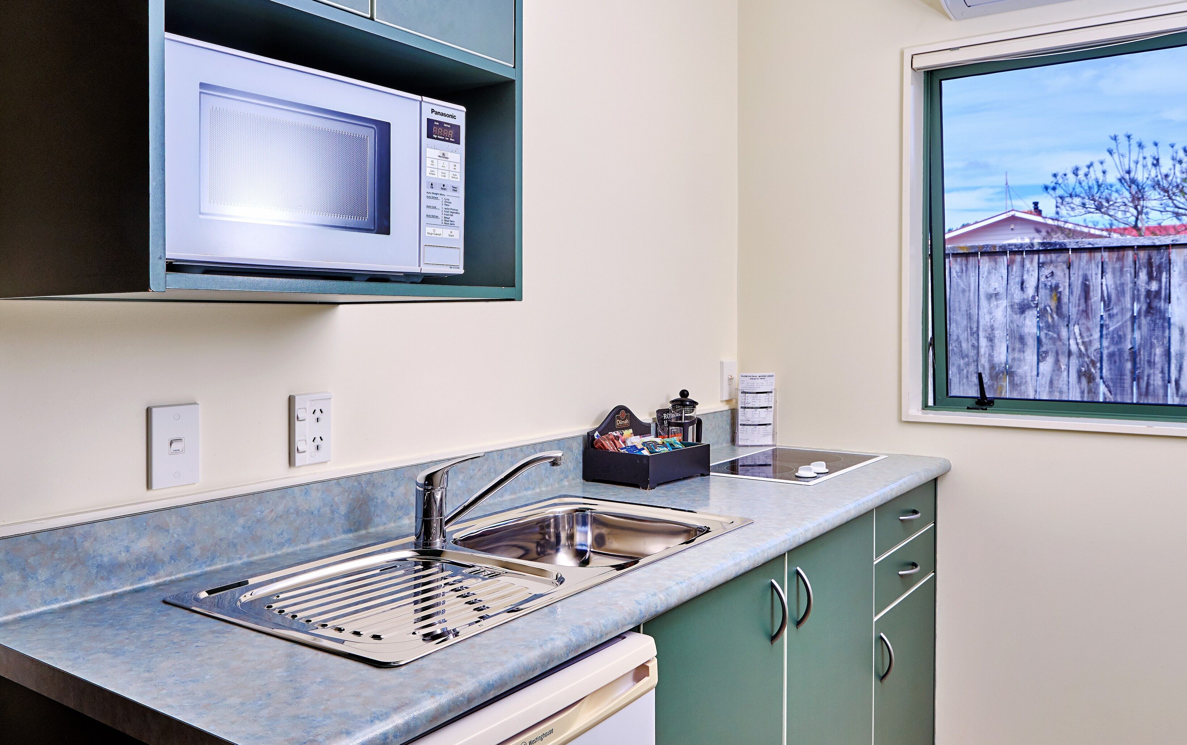 A compact kitchen with a built-in microwave, stainless steel sink, and a window with a view of a wooden fence.