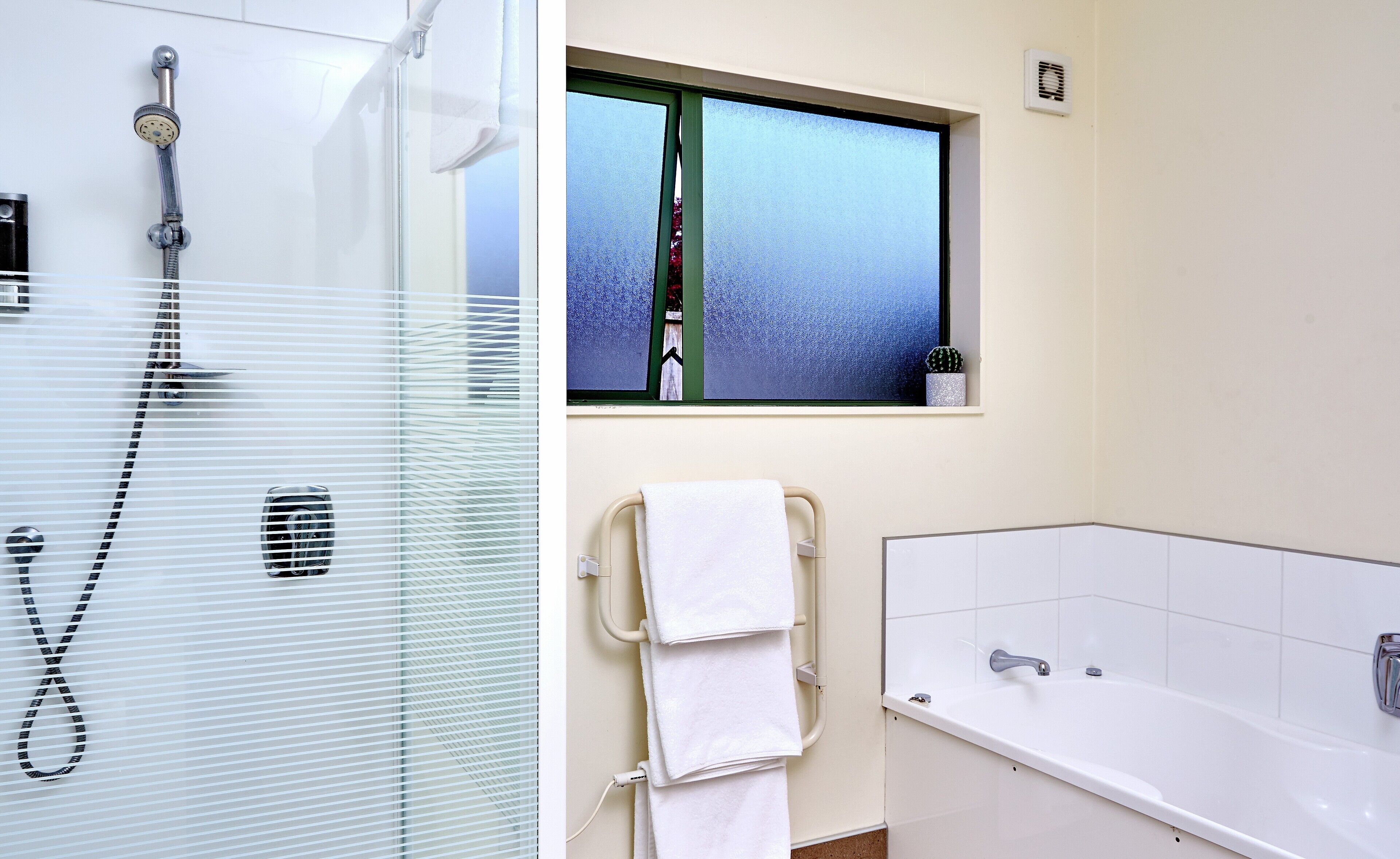 A bathroom with a glass-enclosed shower, a window with frosted glass, a towel rack with two towels, and a white bathtub.