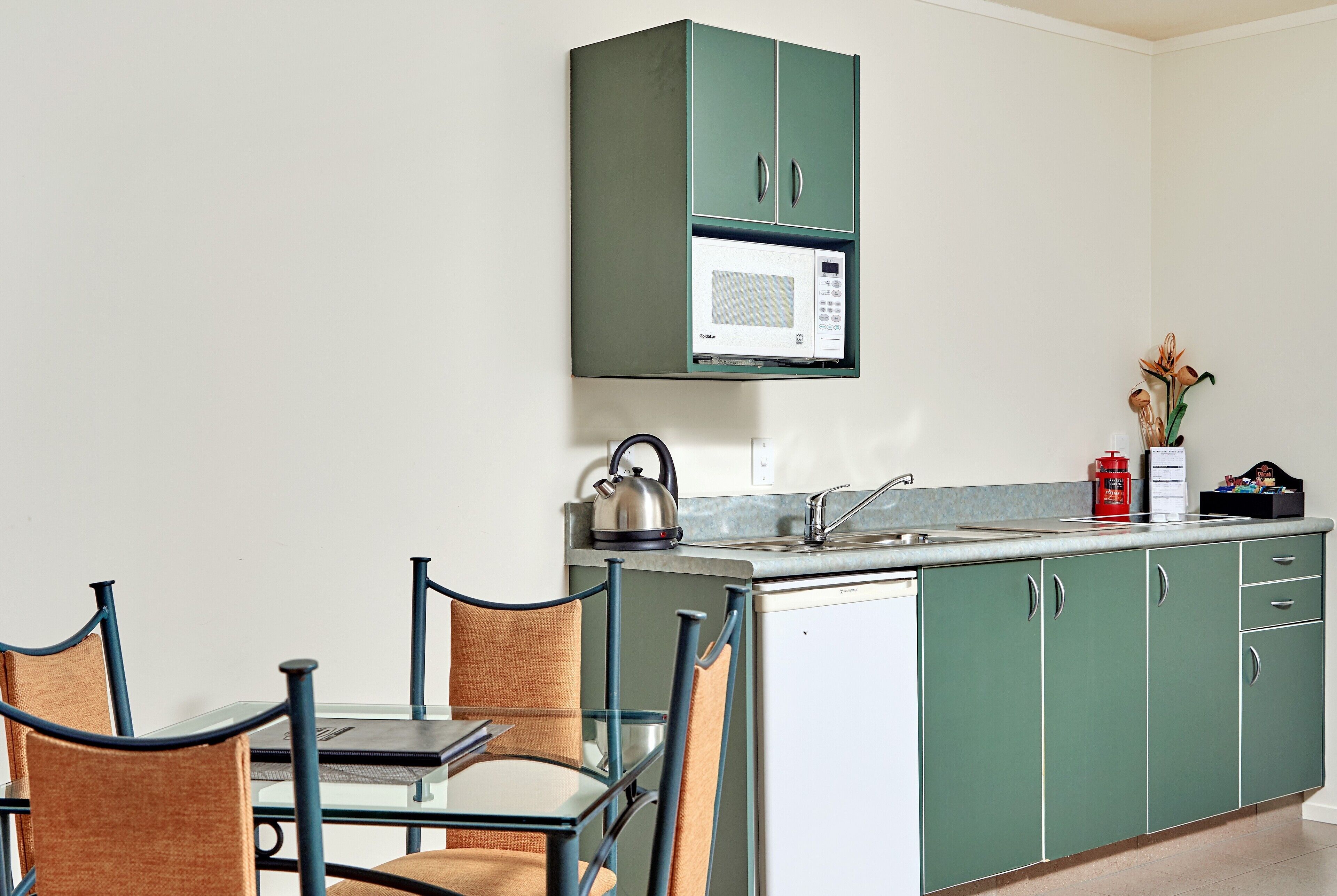 Une cuisine compacte avec des meubles de couleur verte, un four à micro-ondes, une bouilloire et une petite table à manger avec des chaises.